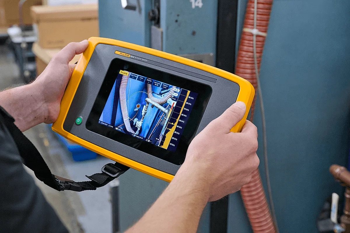 Technician scanning overhead compressed-air piping with a Fluke acoustic imager, the SoundMap overlay highlighting an ultrasonic leak signature