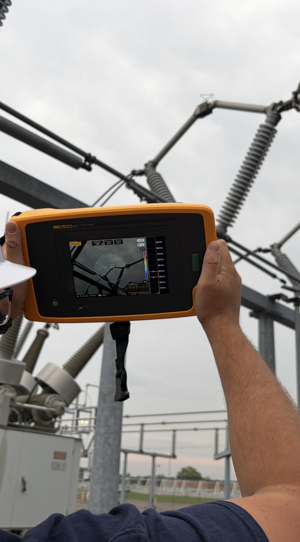 Close-up of a Fluke ii910 acoustic imager screen held overhead, showing the PDQ Mode overlay pinpointing a partial discharge source on an energized substation switch with the frequency scale alongside