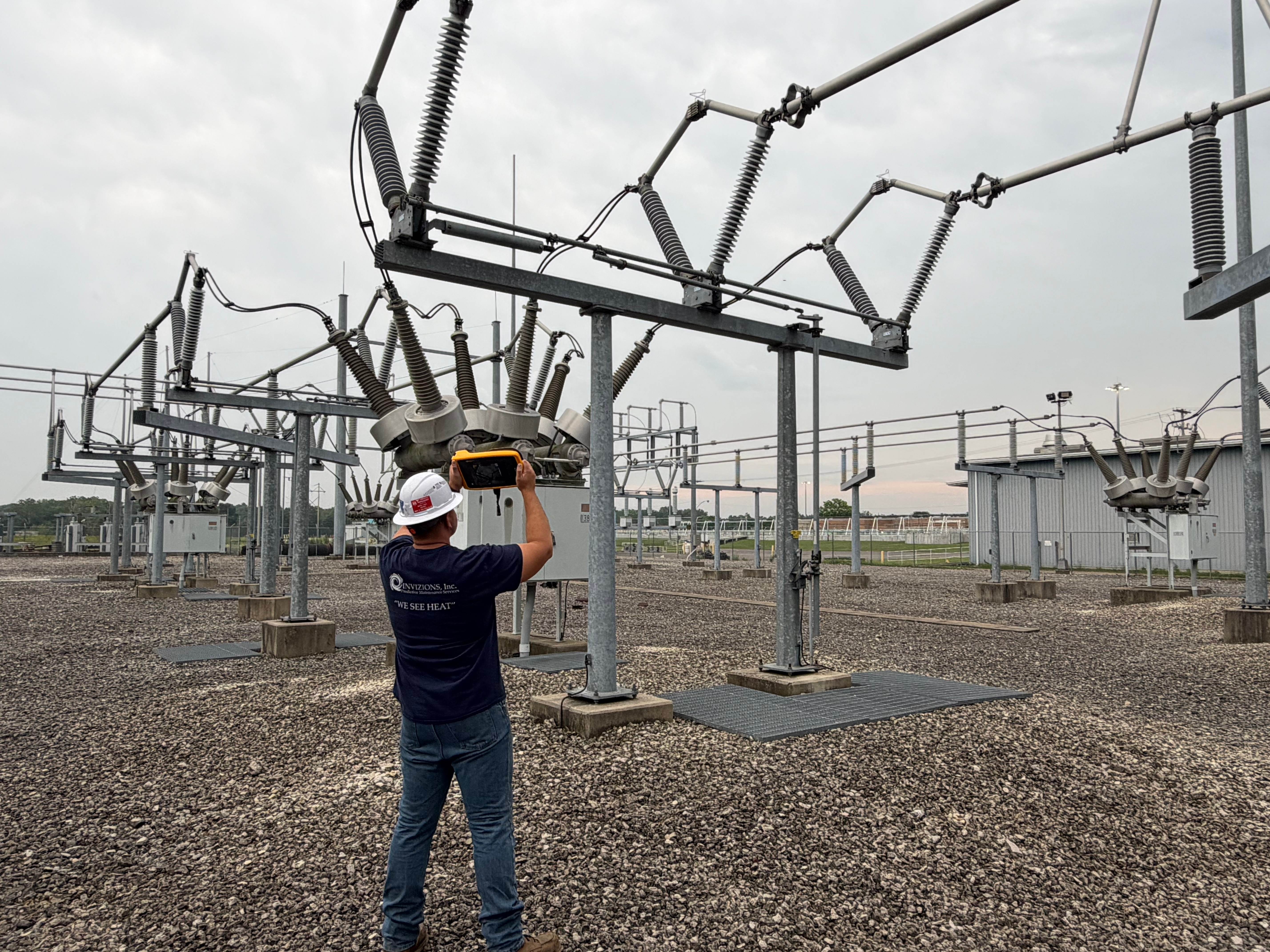 Invizions technician in a hard hat aiming a handheld Fluke acoustic imager at energized 138 kV switchgear during an outdoor substation partial discharge survey